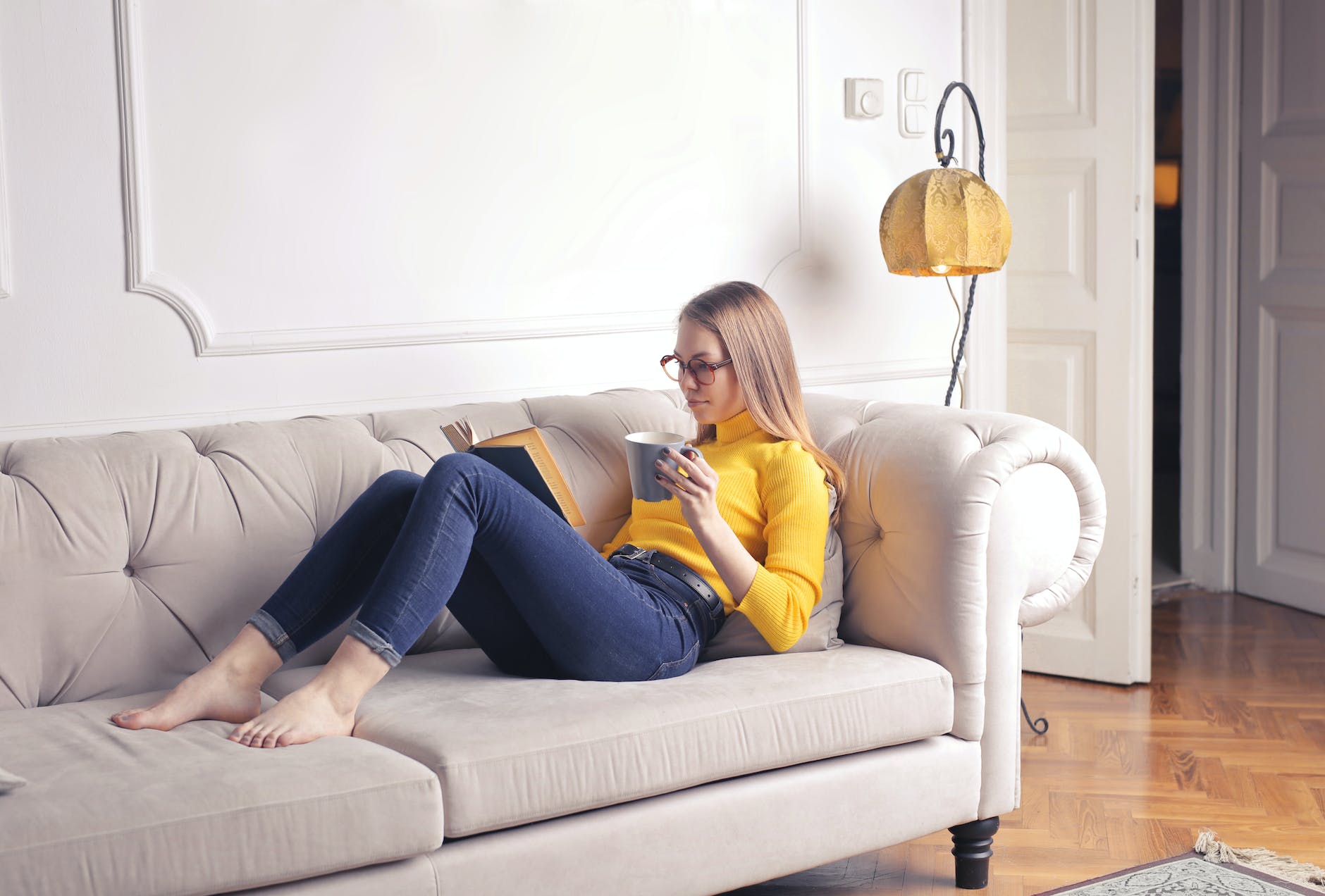 woman sitting on white couch while reading a book, enjoying her day off by doing something for self care